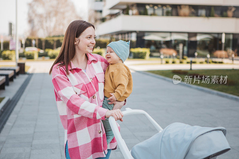 年轻的妈妈抱着婴儿，推着现代的婴儿车，漫步在城市中