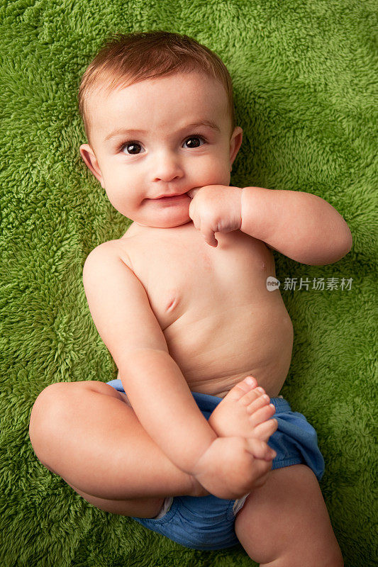 小男孩躺在绿毯子上，嘴里含着手指