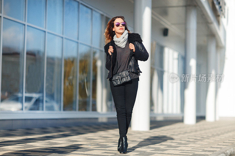 戴着太阳眼镜的女人，黑色皮夹克，黑色牛仔裤在镜子前摆姿势。女性时尚的概念。户外。