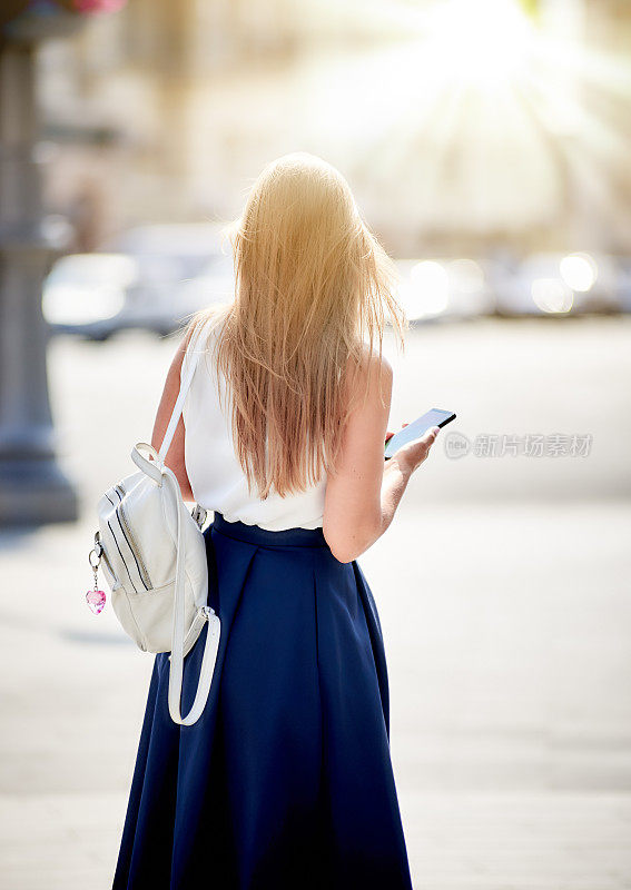 年轻女子背着双肩包，在户外城市街道上行走，使用手机，后视镜