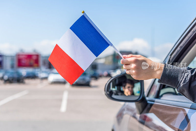 在购物中心的停车场上，一个男孩从打开的车窗里举着法国国旗。概念