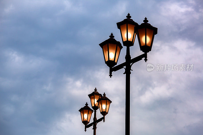 维多利亚风格的灯柱柱户外灯3灯头和透明的玻璃面板，三照明与温暖的辉光电灯在傍晚时间的背景天空与云。