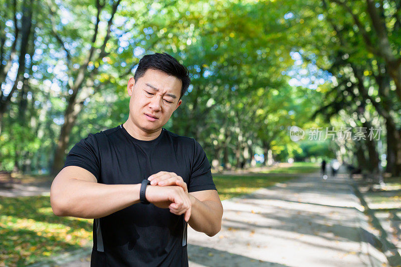 不开心和沮丧的男运动员看着健身与智能手表，糟糕的锻炼结果，亚洲男子在秋天公园的一天锻炼