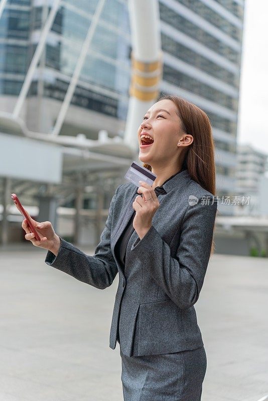 一名年轻的亚洲女商人穿着灰色的办公套装，在商业区用她的智能手机使用信用卡进行在线交易，她微笑着自信