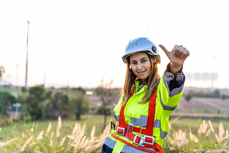 在风力涡轮机农场，穿着制服的白人女工程师微笑着竖起大拇指，庆祝项目成功。快乐工作者享受工作