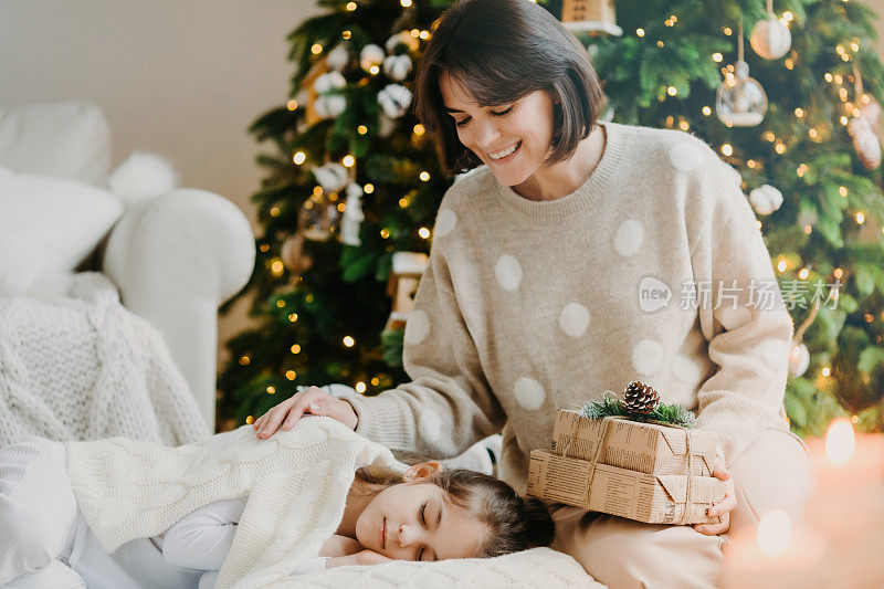 我有个小秘密要告诉你。可爱的母女在家里庆祝新年。妈妈在熟睡的小女孩身边摆姿势，手里拿着礼物，映衬着装饰好的圣诞树。快乐寒假概念