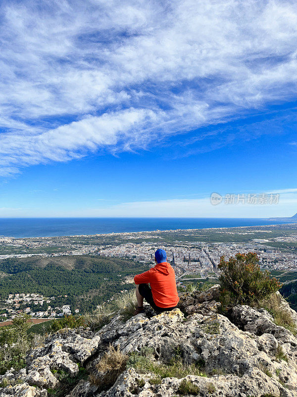 年轻的男性徒步旅行者在爬山后，坐在山顶上，看到包括地中海在内的所有风景，享受着乐趣和放松。