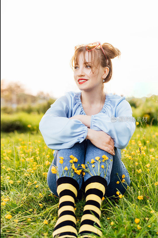 一名年轻女子穿着蓝色上衣、牛仔裤和黑黄相间的条纹袜子，袜子里插着鲜花，坐在盛开的草地上。动画风格。蜜蜂保护概念，开花季节。