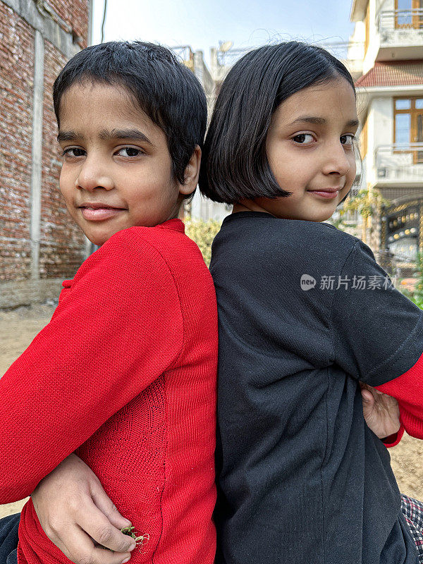 两个年轻的印度女孩在户外摆姿势拍照的特写，姐妹们背靠背坐着，看着相机，前景的焦点，侧面视图