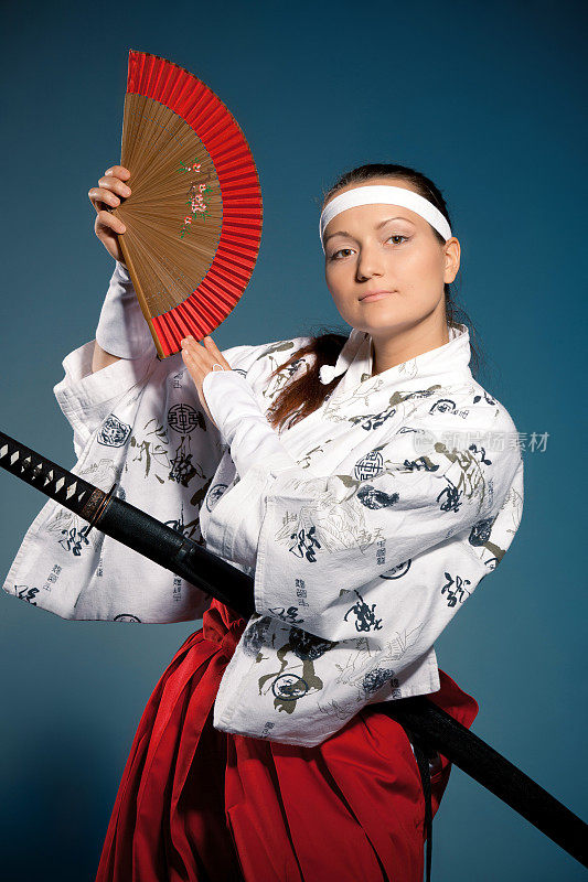 年轻女子在日本衣服与武士刀和扇子摆姿势