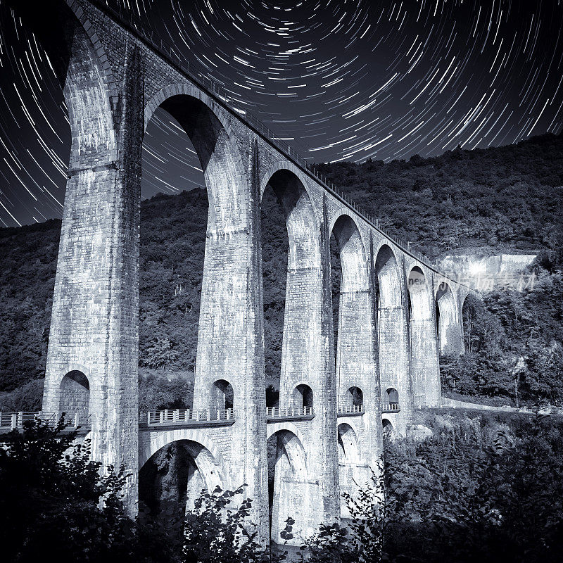 月光下的法国古老的石砌拱桥建筑，夏日夜空中点缀着星光小径，色调为蓝色单色