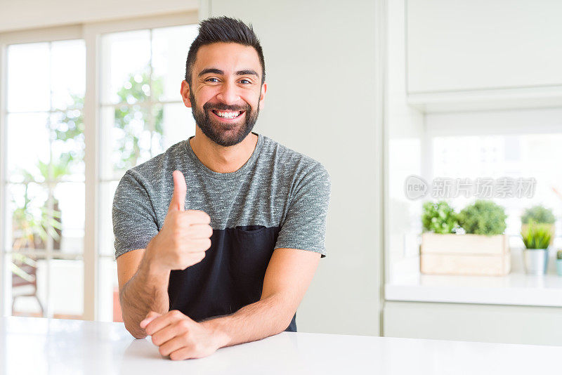 英俊的西班牙男子穿着休闲t恤在家做快乐的拇指手势与手。赞许的表情看着镜头，表示成功。