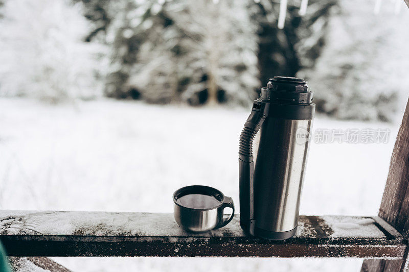 在一所乡村别墅的阳台上，放着热茶的保温瓶，背景是冬天美丽的风景。在白雪覆盖的户外公园里野餐。圣诞节期间的家庭假日