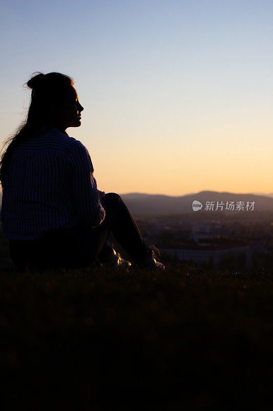 一个女人的黑色剪影坐在草山上俯瞰风景。