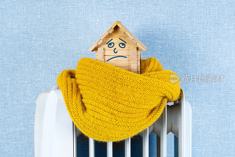 冬天房子里很冷的概念。弱热效率的住宅建筑理念。用玩具木做的房子，用羊毛围巾裹着悲伤的脸，站在一个电暖器上。下雪了。