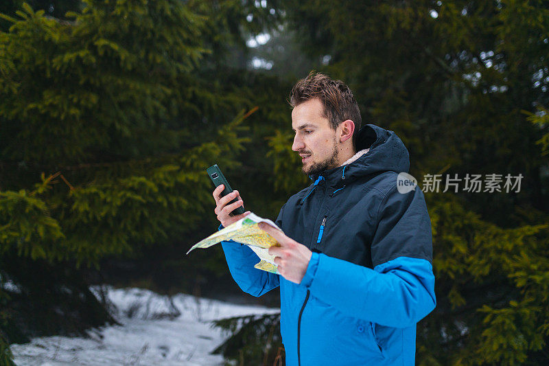 年轻的男性徒步旅行者穿着蓝色夹克，在雾蒙蒙的雪山中迷失了方向，一边看地图，一边看智能手机