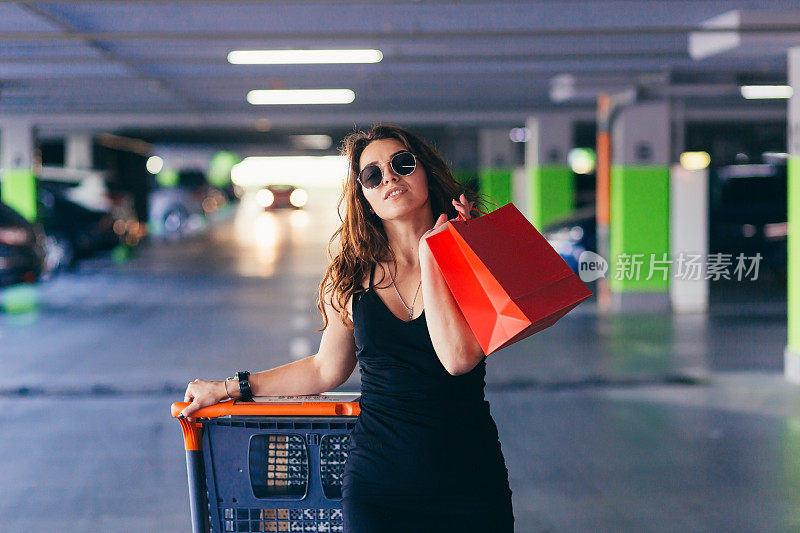年轻漂亮的女子推着购物车，拿着装着商品和物品的包裹，来到购物中心地下停车场的汽车前
