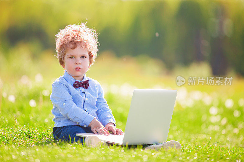 可爱的卷发小男孩打着领结，带着笔记本电脑坐在绿色的草地上。编写计算机软件的神童。