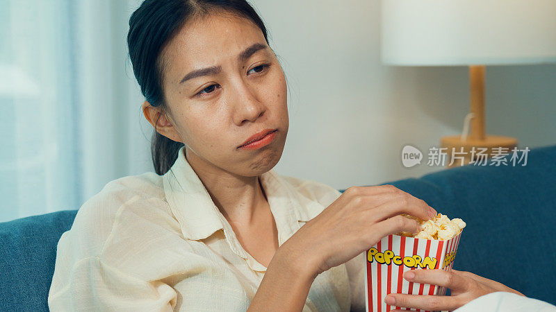 年轻的亚洲好奇女孩在沙发上吃爆米花兴奋地看纪录片频道电影或电视上的悬疑节目在流媒体在线点播，在家里。