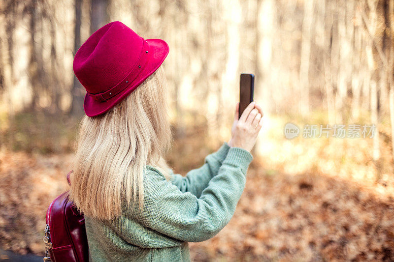 背影上一个女孩戴着红帽子，背着背包拿着手机，拍摄着美妙的秋景。