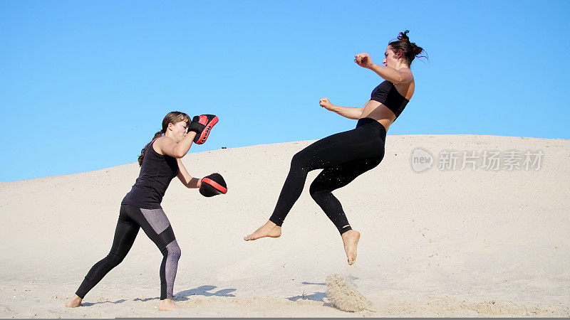 夏天，在烈日炎炎下，两个穿着黑色健身服的年轻女子在荒芜的海滩上，踢着脚，训练着打架。慢动作