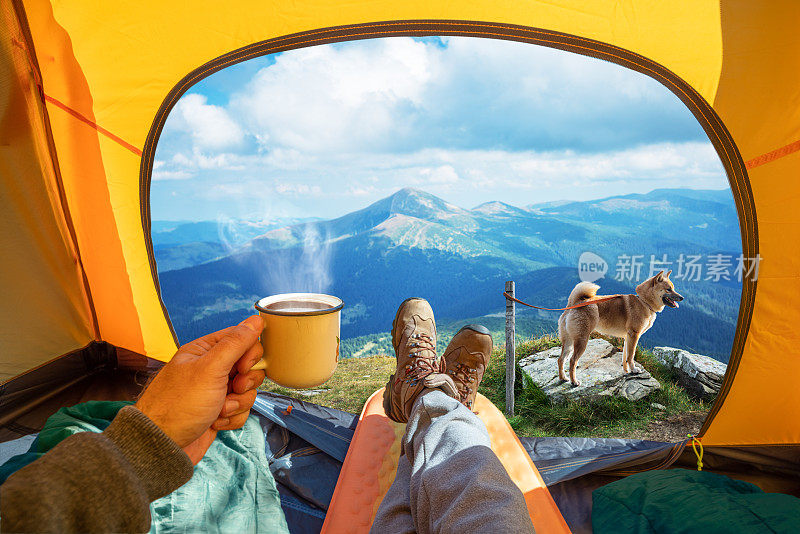 手里拿着一杯热饮，透过敞开的帐篷入口，山顶的美景尽收眼底。在狗的陪伴下，浪漫的徒步旅行和露营的美丽。