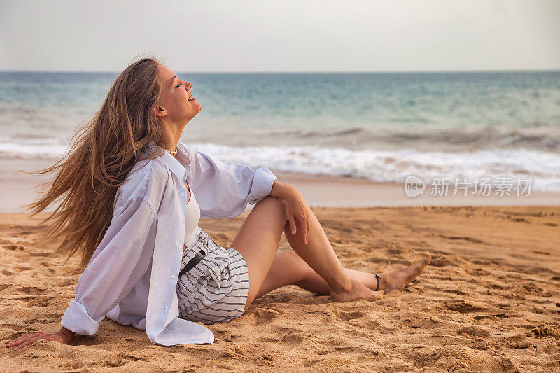 快乐的年轻时尚的女人坐在沙滩上，在大海的背景，看向别处。轻松愉快的热带海洋假期。旅游度假度假概念。复制广告文字空间