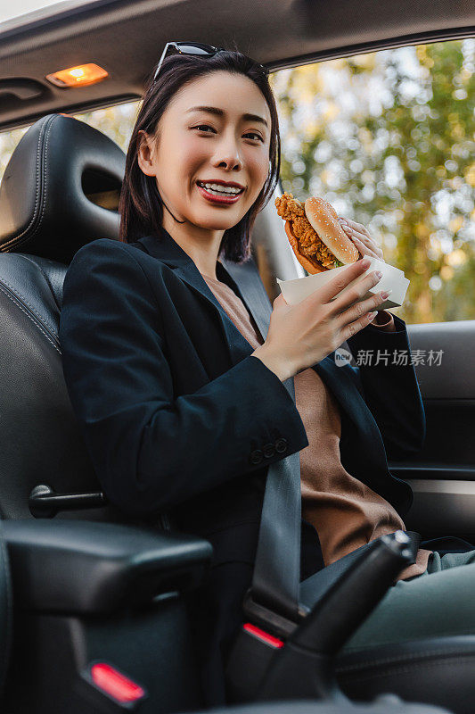 亚洲女人在车里休息，吃汉堡。一个年轻成功快乐的女人正在开车。