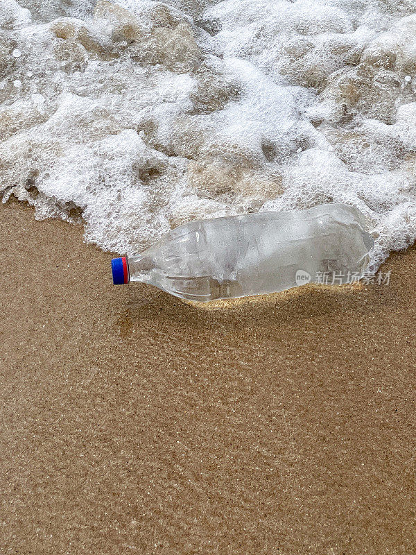 特写图像，空的，透明的塑料饮料瓶，蓝色的盖子被冲到沙滩上，在退潮水的边缘，破碎的海浪冲刷海洋垃圾和岸上的污染，乱扔的沙子，肮脏的海滩，关注前景，复制空间