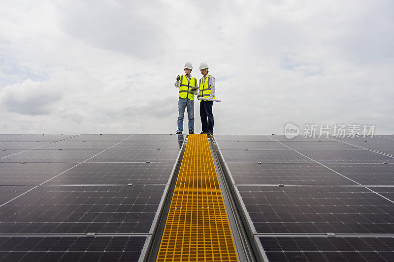 两个年轻的亚洲工程师在工厂屋顶检查太阳能电池板的安装情况，对照电脑中的图纸检查太阳能电池板的安装情况。