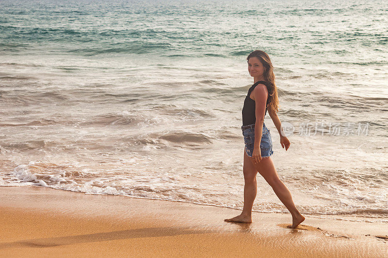 快乐的年轻女子在牛仔短裤走在沙滩上的海浪背景，暑假。旅行者女士走在海边，看着相机。旅游度假概念。复制广告文字空间