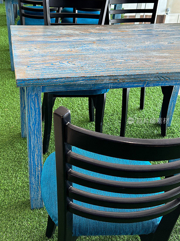 现代餐厅设置的全画幅图像，室内用餐木制桌椅与蓝色垫垫设置在人造草草坪地板，玻璃窗户墙，重点在前景