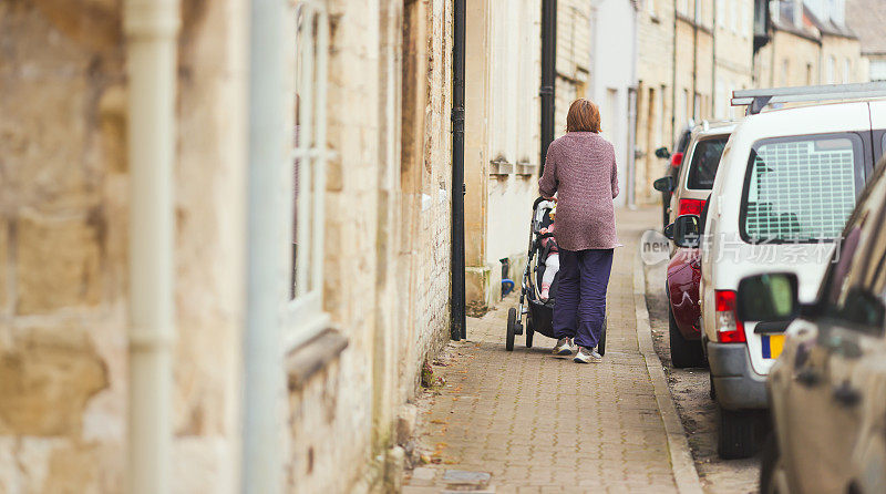 在英国格洛斯特郡的赛伦塞斯特，一位祖母推着她的孙子走在一条小路上，小路周围都是古老的科茨沃尔德石屋，街上停着汽车
