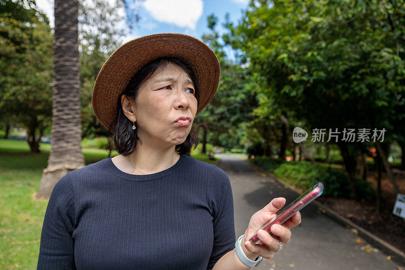 一个亚洲女人在公园里拿着手机，一脸困惑