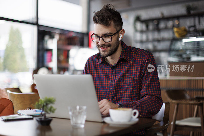 微笑的年轻人戴着耳机在咖啡馆里使用笔记本电脑