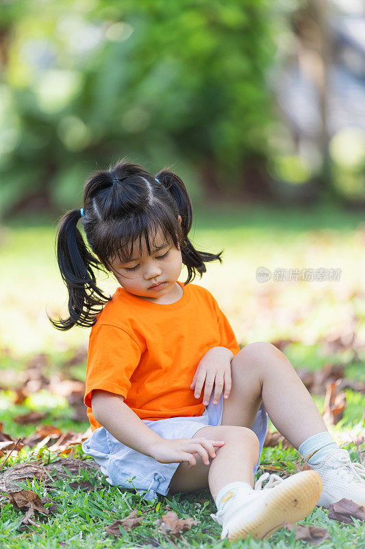 一个穿着橙色衬衫的小女孩坐在草地上。她低头看着自己的腿