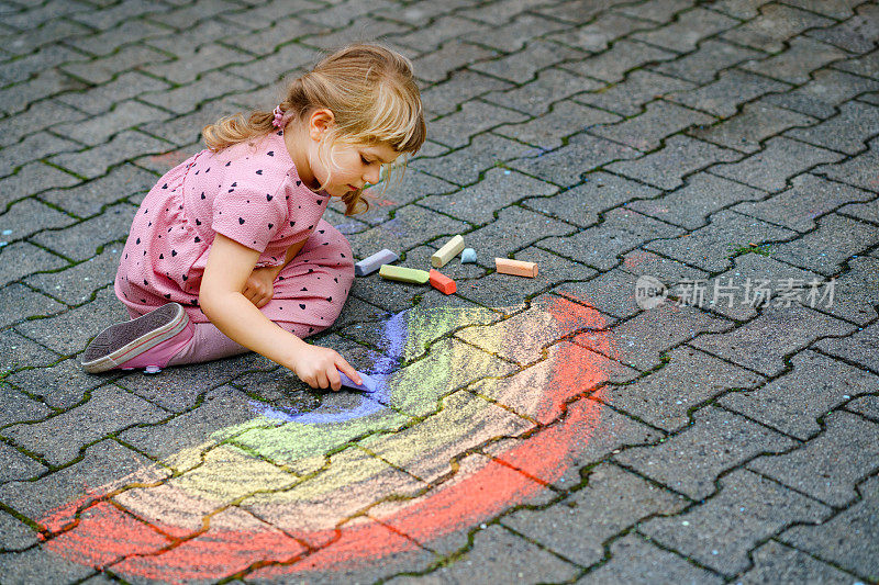 学前班的小女孩用彩色粉笔在后院的地上画彩虹。积极快乐的幼儿绘画和创造图片。夏天有创意的户外活动。