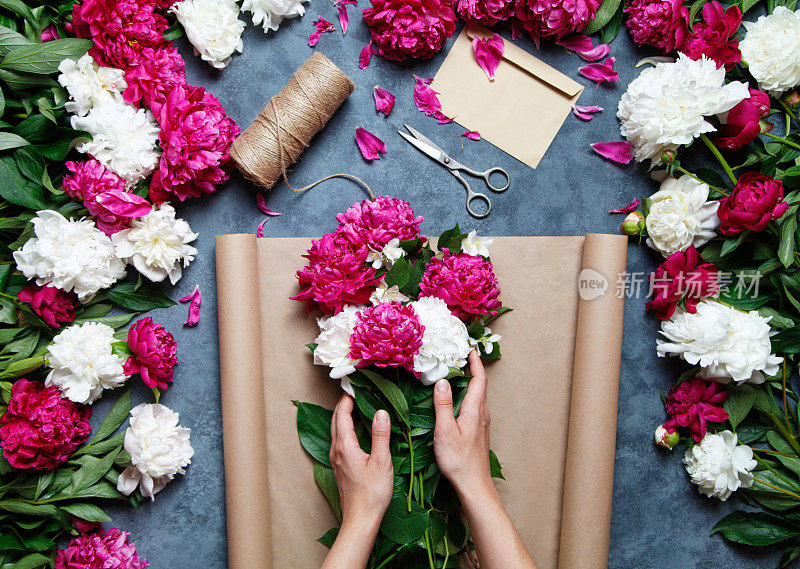 工作中的花商:一个漂亮的女人正在灰色的工作桌上制作夏季的牡丹花束。牛皮纸，剪刀，信封放在桌上。从以上观点。