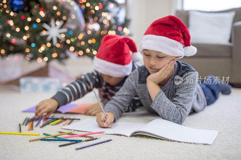 两个小男孩戴着精灵帽躺在圣诞树前写下他们的愿望清单
