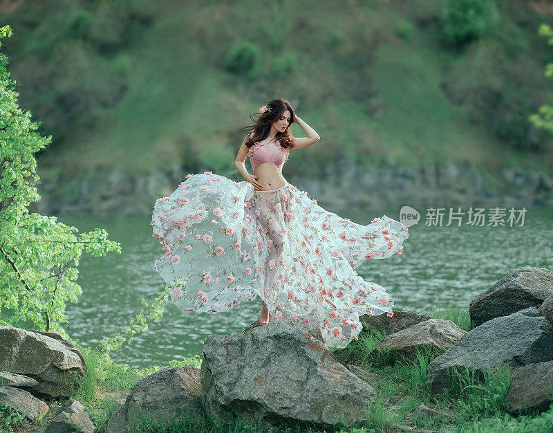 春姑娘挽着波浪形的头发，以山水为背景，翩翩起舞。她穿着一件粉红色的花朵连衣裙，在风中飘动，突出了性感的轮廓。艺术照片