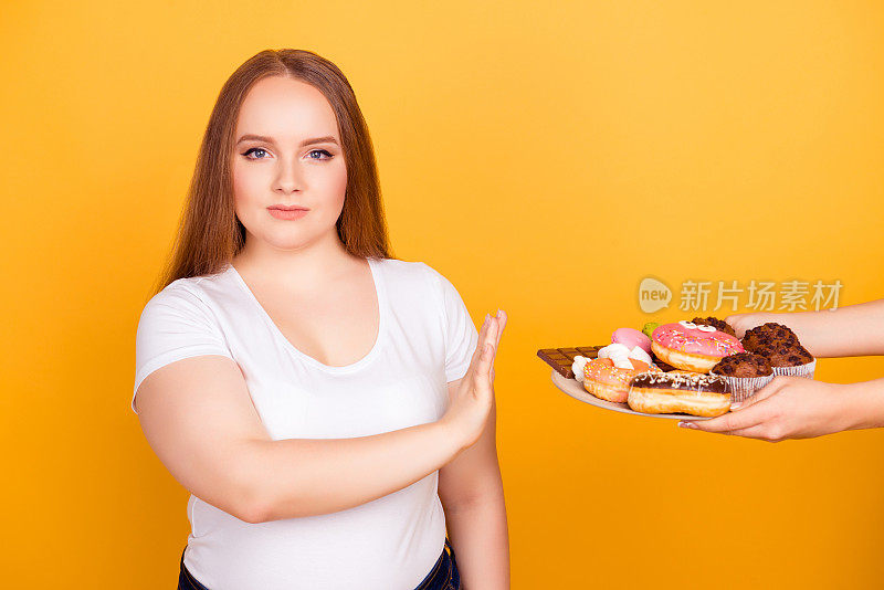 我反对吃含脂肪的食品!一个身穿白色t恤的意志坚强的女人正在拒绝吃盘子上的美味的糖果，孤立在明亮的黄色背景