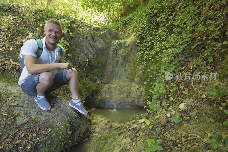 成年徒步旅行者坐在小溪旁，周围长满了绿色植物