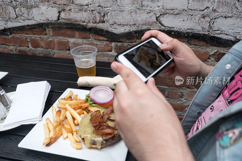 一名男子拿着手机给他的食物拍照。智能手机食品摄影。在户外酒吧用手机给汉堡、啤酒和炸薯条拍照。