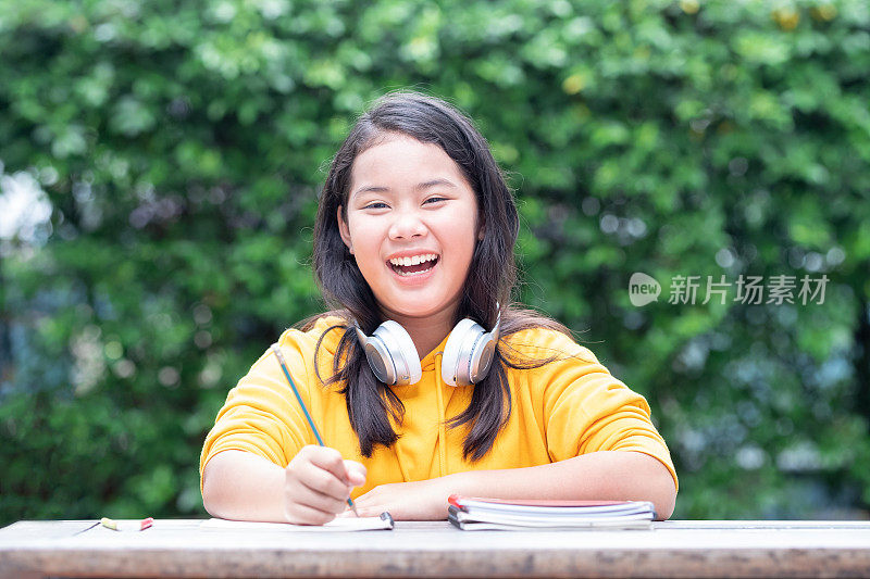 微笑的亚洲小女生在公园里做作业，她的脖子上戴着耳机