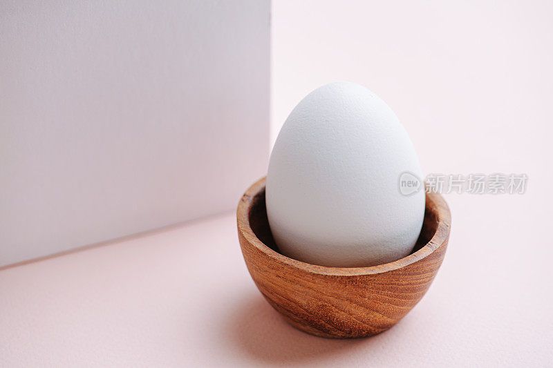 木蛋里的白蛋立在粉红色的表面上，旁边是一个白色的纸立方体