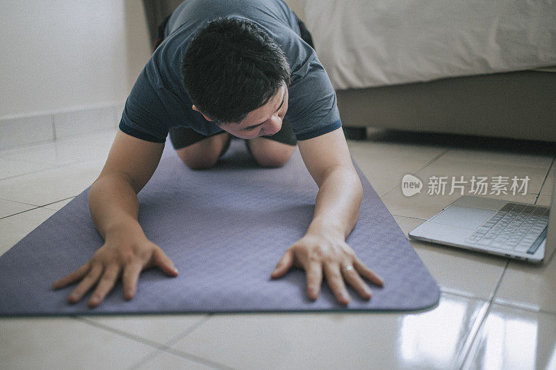 一名亚裔中国中年男子在卧室的瑜伽垫上练习瑜伽，抱着他的笔记本电脑