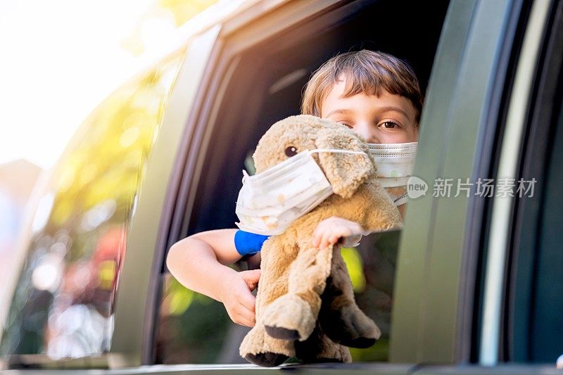 一个悲伤的小男孩戴着保护面罩，抱着一只泰迪熊，看着车窗外的摄像头