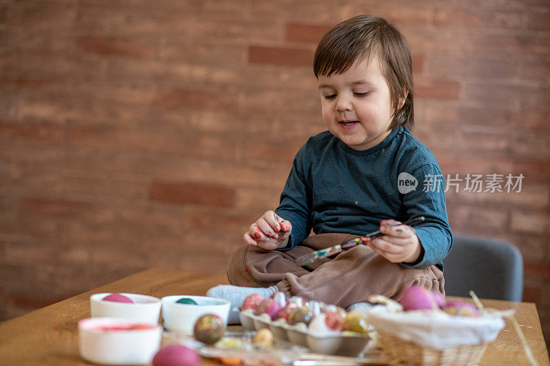 可爱的小男孩正坐在桌子上，用毛笔在复活节彩蛋上画画。