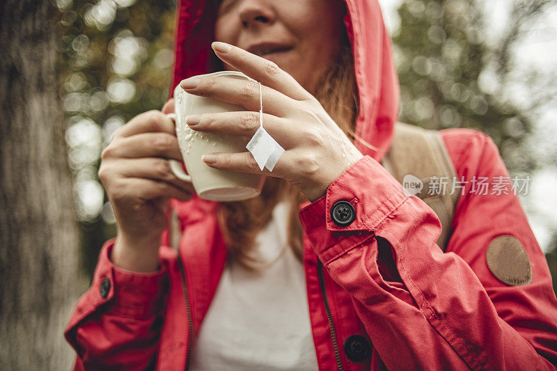 一个年轻的橘黄色头发的女人，穿着红色的雨衣，在雨中走过树林，拿着一杯热茶-放松在一个雨天的自然