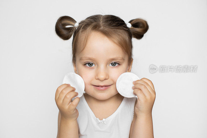 摄影棚拍摄了一个漂亮的小女孩，用化妆棉洗脸，在镜头前摆姿势，面带微笑，享受护肤程序。近距离肖像孤立的白色背景